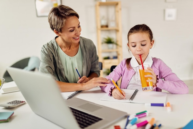 Niña bebiendo jugo mientras hace la tarea con su madre