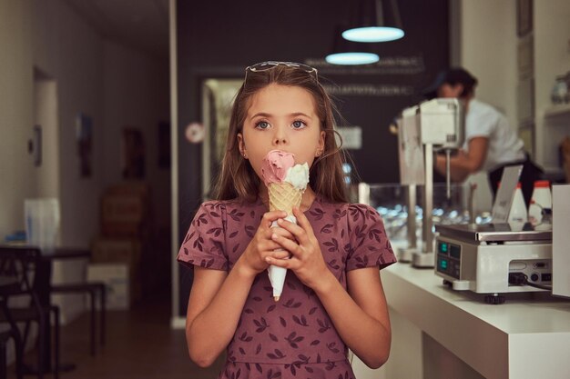 Una niña de belleza con un vestido de moda comiendo fresa, parada en una heladería.