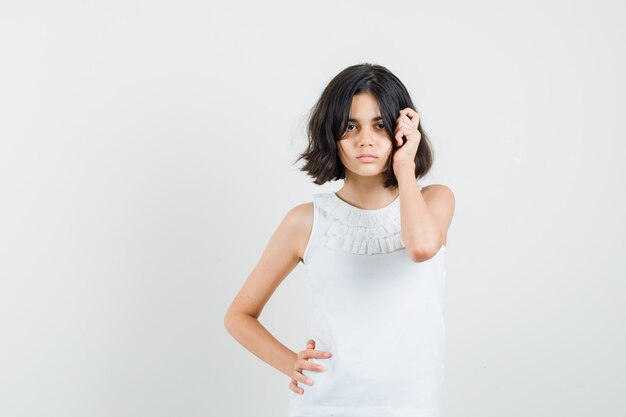 Niña en blusa blanca con la mano en el cabello y aspecto cansado, vista frontal.