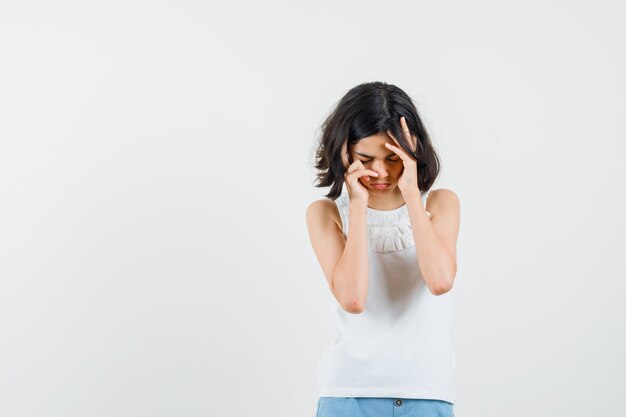 Niña de blusa blanca, pantalones cortos con fuerte dolor de cabeza y aspecto fatigado, vista frontal.