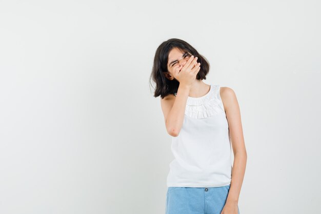 Niña con blusa blanca, pantalones cortos sosteniendo la mano en la boca y mirando feliz, vista frontal.