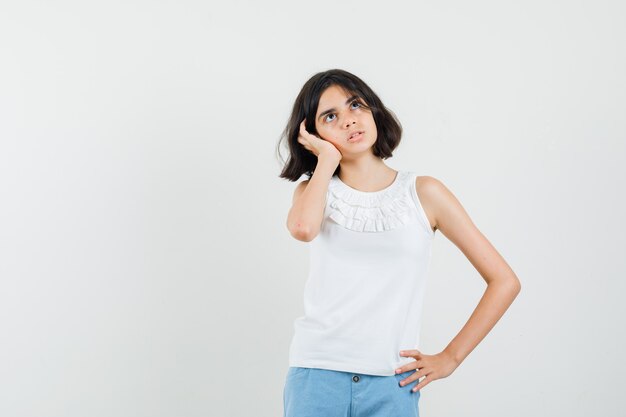 Niña en blusa blanca, pantalones cortos sosteniendo la mano en la oreja y mirando pensativo, vista frontal.