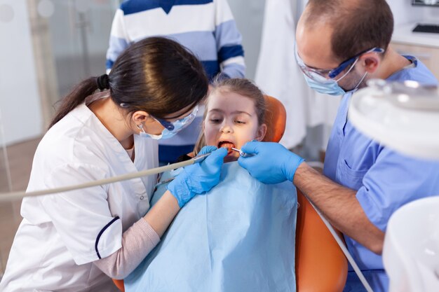 Niña con la boca abierta en el curso del tratamiento de la caries sentado en el sillón dental. Madre con su hijo en la clínica de estomatología para examinar los dientes con instrumentos modernos.