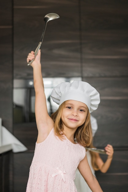 Niña bonita con el brazo levantado sosteniendo la cuchara que lleva el sombrero del cocinero