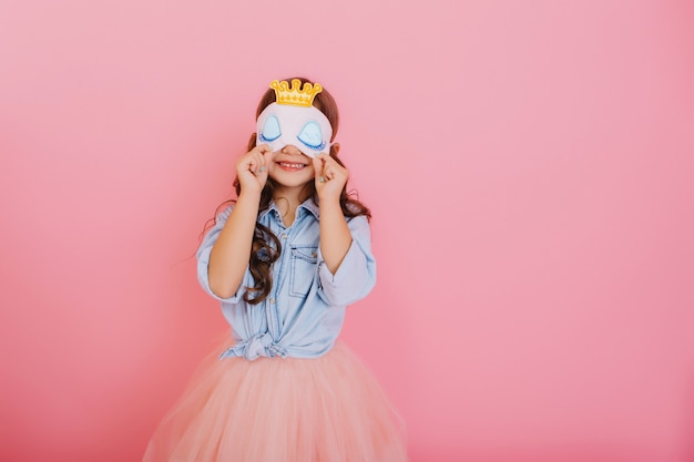 Niña bonita con cabello largo morena en falda de tul con antifaz para dormir con ojos azules y corona dorada aislada sobre fondo rosa. Celebrando la fiesta de cumpleaños, divirtiéndose en el carnaval para niños