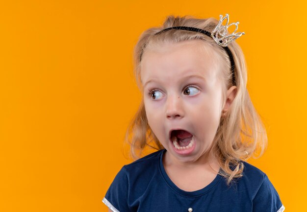 Una niña bonita con camisa azul marino en diadema de corona abriendo la boca y mirando de lado en una pared naranja