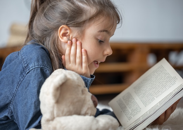 Niña bonita en casa, tirada en el suelo con su juguete favorito y lee el libro.