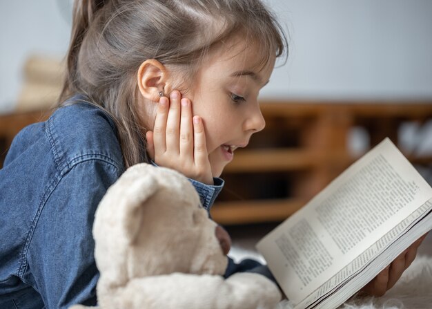 Niña bonita en casa, tirada en el suelo con su juguete favorito y lee el libro.