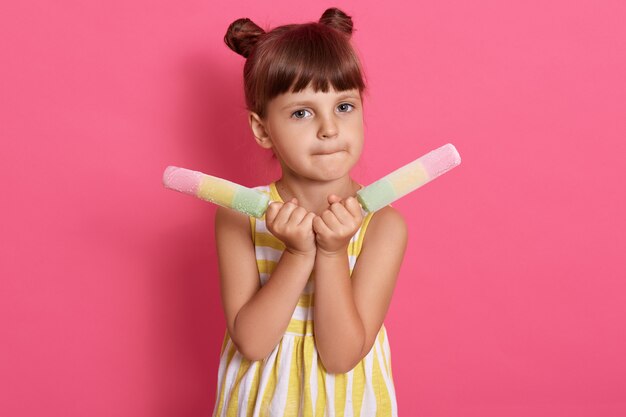 Niña bonita comiendo comiendo dos grandes helados, vestido de blanco y amarillo, con dos bollos de pelo, posando contra la pared de color de rosa con dos sorbetes.