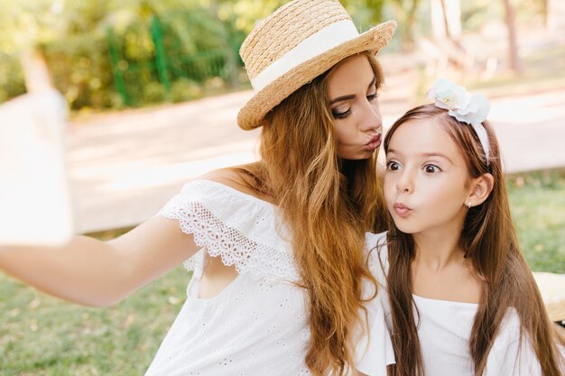 Niña bonita con grandes ojos marrones posando con expresión de cara de sorpresa mientras su madre sostiene el teléfono inteligente. Mujer elegante besando a su hija en la frente y haciendo selfie.
