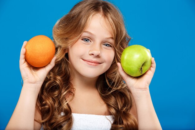 Niña bonita con manzana y naranja sobre pared azul