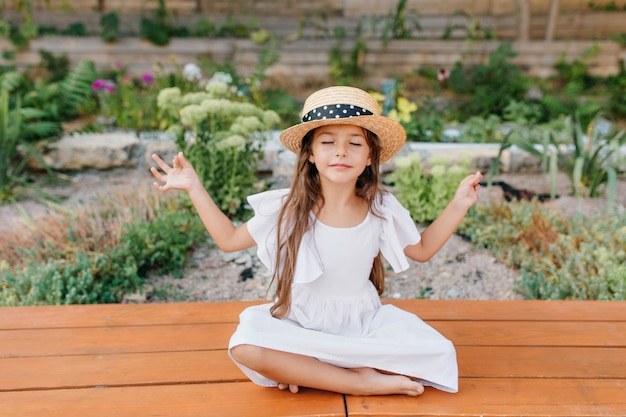 Niña bonita morena con sombrero de paja sentado cerca del macizo de flores en posición de loto con los ojos cerrados. Niña con vestido blanco haciendo yoga en el jardín