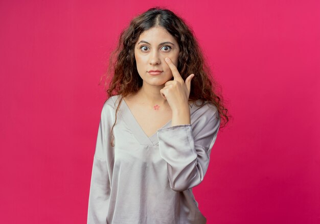 Niña bonita poniendo el dedo en el ojo aislado en la pared rosa