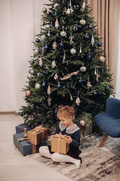 La niña bonita se sienta debajo de un gran árbol de navidad, sostiene su presente y sonríe