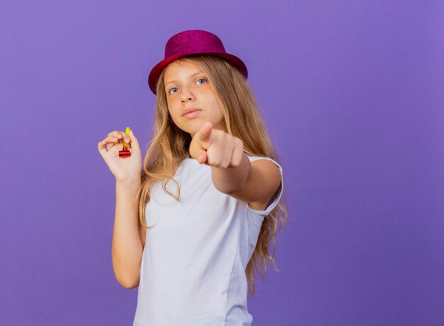 Niña bonita con sombrero de fiesta apuntando con el dedo índice a la cámara con cara seria, concepto de fiesta de cumpleaños de pie sobre fondo púrpura