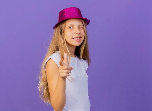 Niña bonita con sombrero de fiesta apuntando con el dedo índice a la cámara sonriendo y guiñando un ojo, concepto de fiesta de cumpleaños de pie sobre fondo púrpura