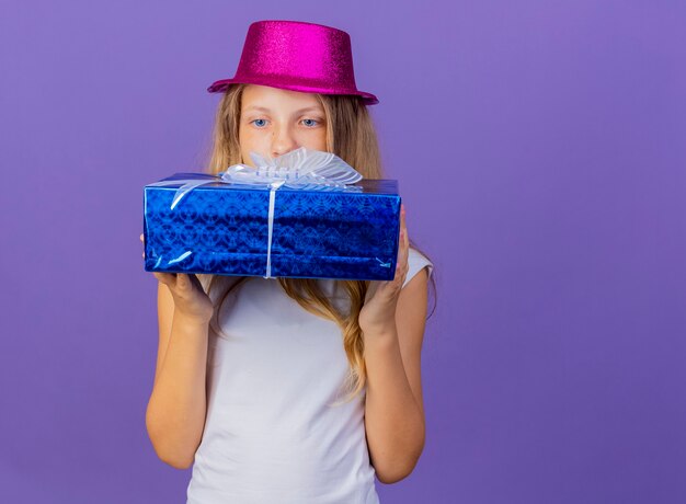 Niña bonita con sombrero de fiesta con caja de regalo mirando intrigado, concepto de fiesta de cumpleaños de pie sobre fondo púrpura