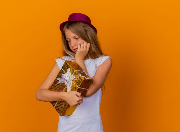 Niña bonita en sombrero de fiesta con caja de regalo mirando a un lado con expresión pensativa pensando, concepto de fiesta de cumpleaños de pie sobre fondo naranja