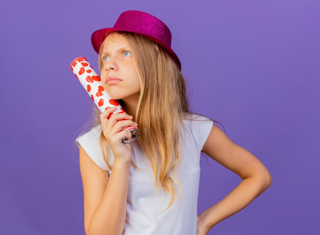 Niña bonita con sombrero de fiesta celebración galleta de fiesta mirando desconcertado, concepto de fiesta de cumpleaños de pie sobre fondo púrpura