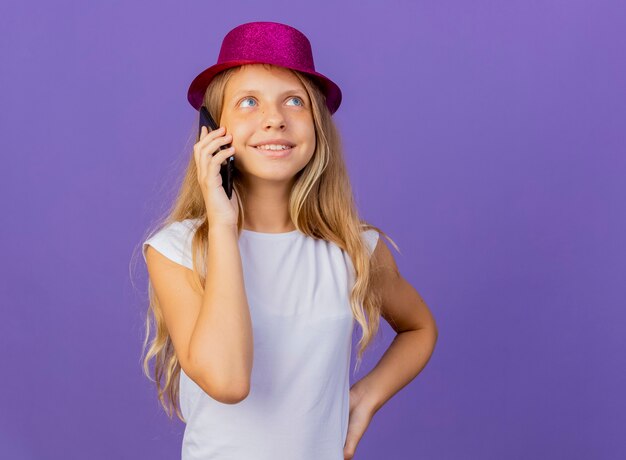 Niña bonita con sombrero de fiesta hablando por teléfono móvil sonriendo, concepto de fiesta de cumpleaños de pie sobre fondo púrpura