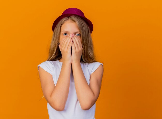 Niña bonita con sombrero de fiesta mirando a la cámara conmocionada cubriendo la boca con las manos, concepto de fiesta de cumpleaños de pie sobre fondo naranja