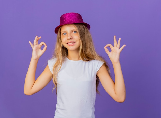 Niña bonita con sombrero de fiesta mirando a la cámara sonriendo con cara feliz mostrando ok cantar, concepto de fiesta de cumpleaños de pie sobre fondo púrpura