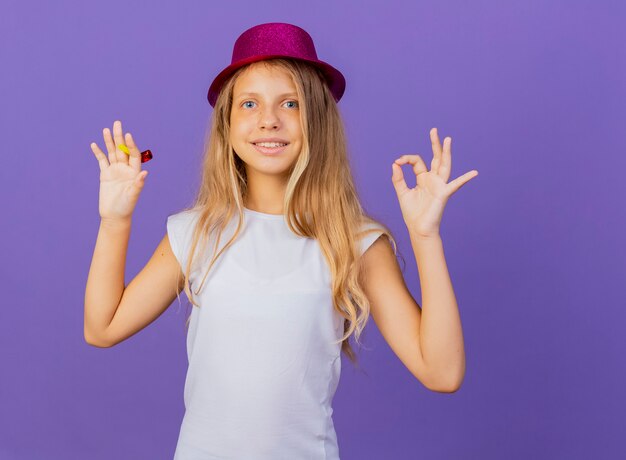 Niña bonita con sombrero de fiesta mirando a la cámara sonriendo mostrando signo de ok, concepto de fiesta de cumpleaños de pie sobre fondo púrpura