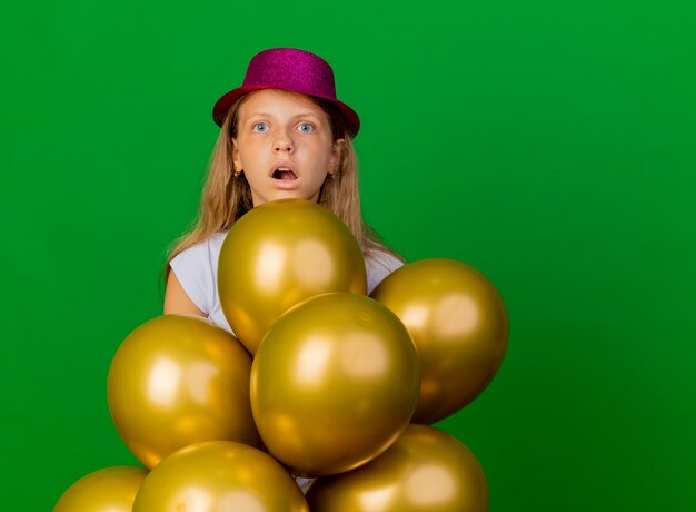 Niña bonita con sombrero de fiesta con montón de globos mirando a un lado asombrado y sorprendido, concepto de fiesta de cumpleaños de pie sobre fondo verde