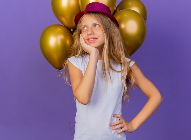 Niña bonita con sombrero de fiesta con montón de globos mirando a un lado con cara feliz sintiendo emociones positivas sonriendo, concepto de fiesta de cumpleaños de pie sobre fondo púrpura