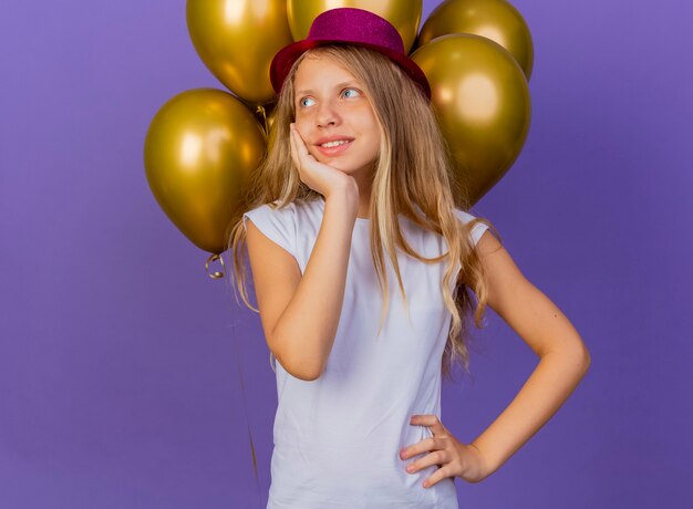 Niña bonita con sombrero de fiesta con montón de globos mirando a un lado con cara feliz sintiendo emociones positivas sonriendo, concepto de fiesta de cumpleaños de pie sobre fondo púrpura