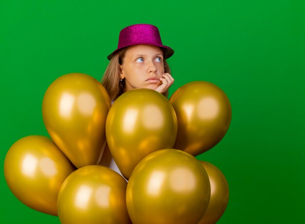 Niña bonita en sombrero de fiesta con montón de globos mirando a un lado desconcertado, concepto de fiesta de cumpleaños de pie sobre fondo verde