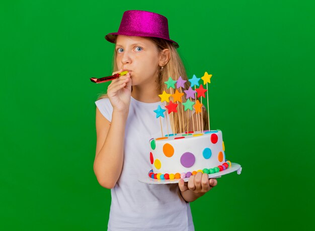 Niña bonita en sombrero de fiesta con pastel de cumpleaños que sopla el silbato