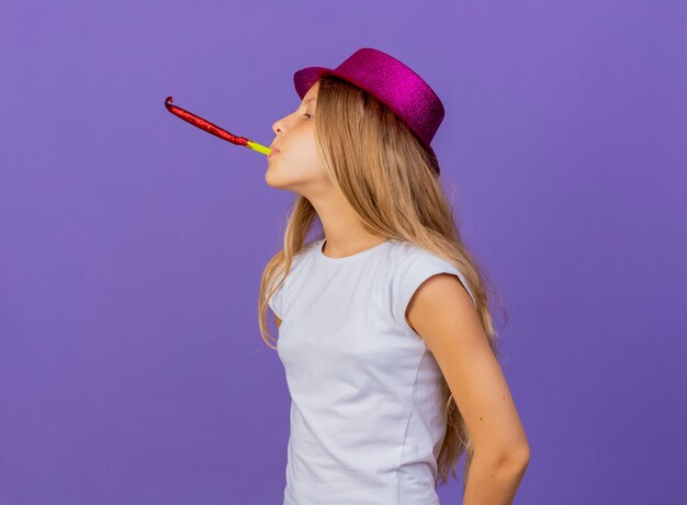 Niña bonita con sombrero de fiesta que sopla el silbato, concepto de fiesta de cumpleaños de pie sobre fondo púrpura