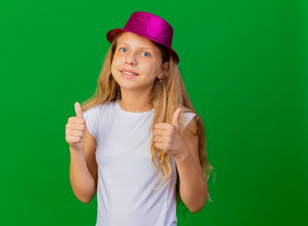 Niña bonita con sombrero de fiesta sonriendo mostrando los pulgares para arriba