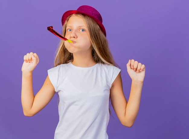 Foto gratuita niña bonita en sombrero de fiesta soplando silbato apretando los puños feliz y emocionado, concepto de fiesta de cumpleaños de pie sobre fondo púrpura