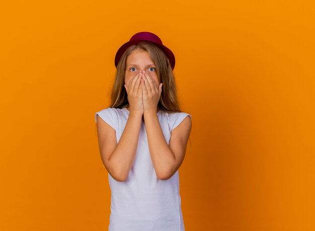 Niña bonita con sombrero de fiesta se sorprendió cubriendo la boca con las manos, concepto de fiesta de cumpleaños