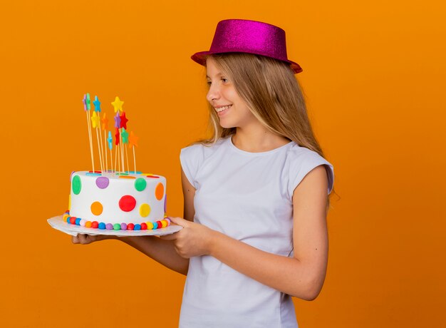 Niña bonita con sombrero de fiesta sosteniendo pastel de cumpleaños mirando a un lado con una sonrisa en la cara, concepto de fiesta de cumpleaños de pie sobre fondo naranja