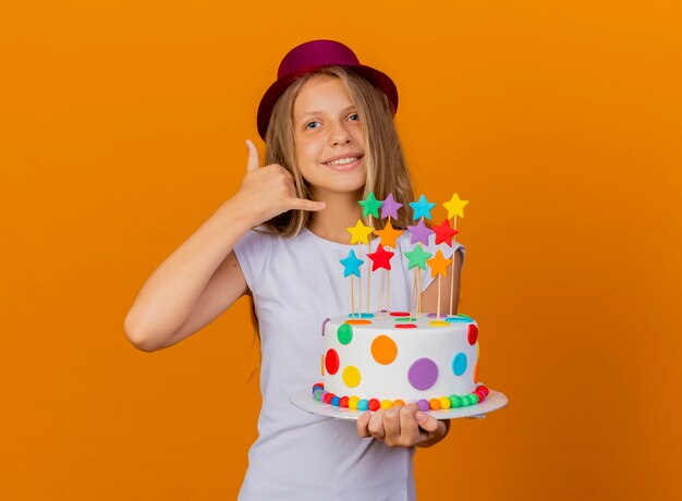 Niña bonita con sombrero de fiesta sosteniendo pastel de cumpleaños sonriendo haciendo gesto de llamarme, concepto de fiesta de cumpleaños