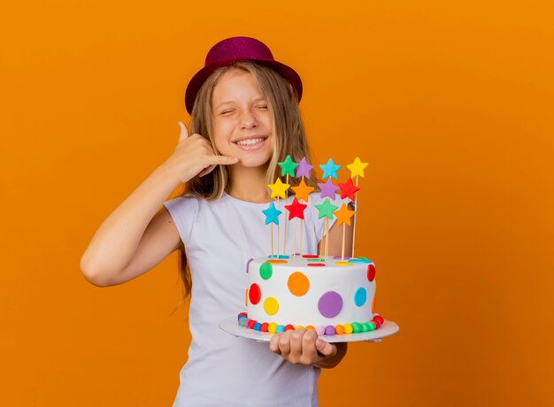 Niña bonita con sombrero de fiesta sosteniendo pastel de cumpleaños sonriendo haciendo gesto de llamarme, concepto de fiesta de cumpleaños