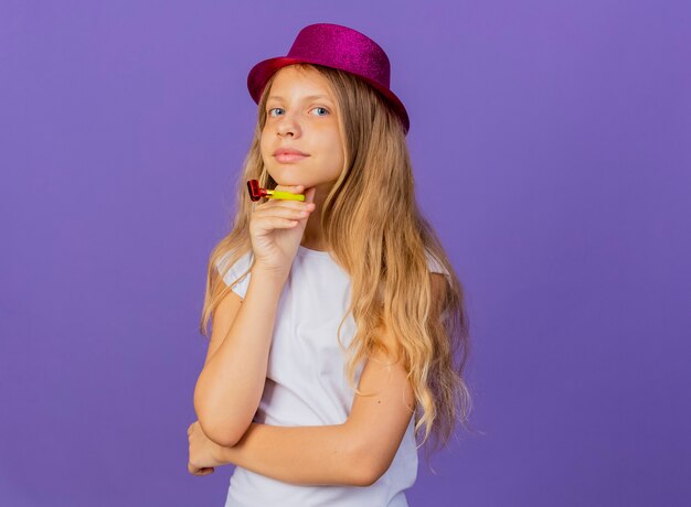 Niña bonita en sombrero de fiesta sosteniendo silbato mirando a la cámara sonriendo con cara feliz, concepto de fiesta de cumpleaños de pie sobre fondo púrpura