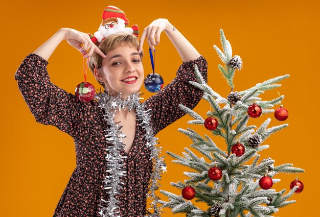 Niña bonita sonriente con diadema de Papá Noel y guirnalda de oropel alrededor del cuello de pie cerca del árbol de Navidad decorado sosteniendo adorno de Navidad cerca de la cabeza aislada en la pared naranja