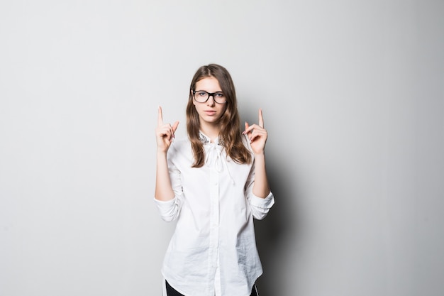 Niña bonita sonriente con gafas vestidas con una estricta camiseta blanca de oficina se encuentra frente a la pared blanca