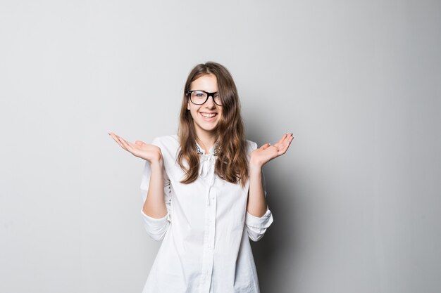 Niña bonita sonriente con gafas vestidas con una estricta camiseta blanca de oficina se encuentra frente a la pared blanca