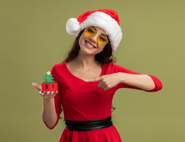 Niña bonita sonriente con gorro de Papá Noel y gafas sosteniendo y apuntando al juguete del árbol de Navidad con fecha aislado en la pared verde oliva