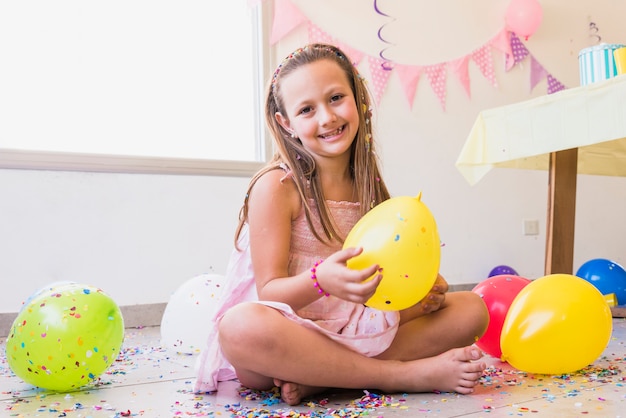 Niña bonita sonriente que se sienta en piso con confeti y globos