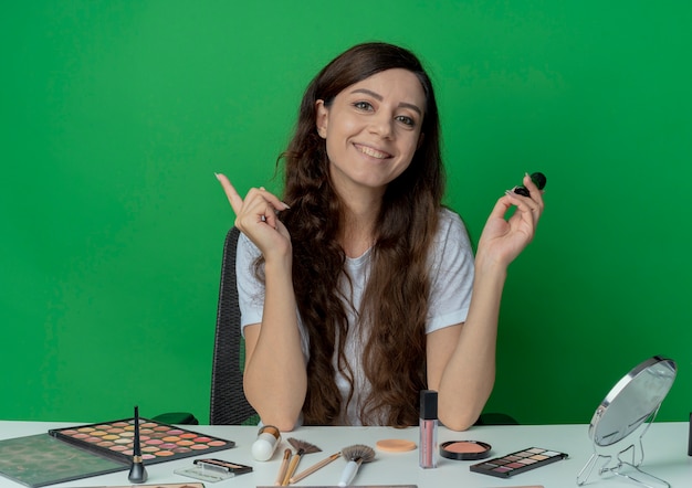 Niña bonita sonriente sentada en la mesa de maquillaje con herramientas de maquillaje sosteniendo pincel de maquillaje y levantando el dedo aislado sobre fondo verde