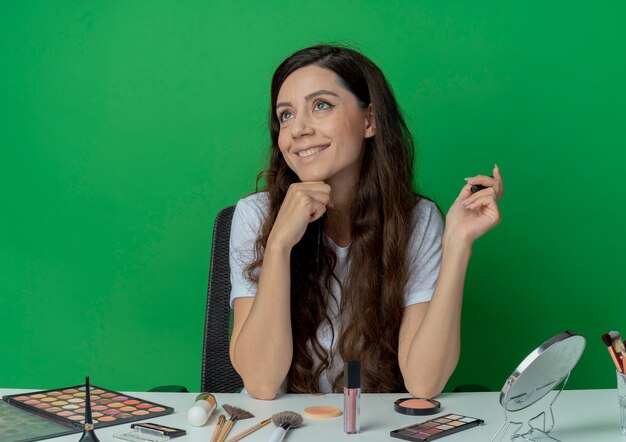 Niña bonita sonriente sentada en la mesa de maquillaje con herramientas de maquillaje sosteniendo rímel tocando la barbilla y mirando al lado aislado sobre fondo verde