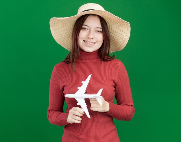 Niña bonita sonriente con sombrero de playa sosteniendo avión modelo aislado en la pared verde con espacio de copia