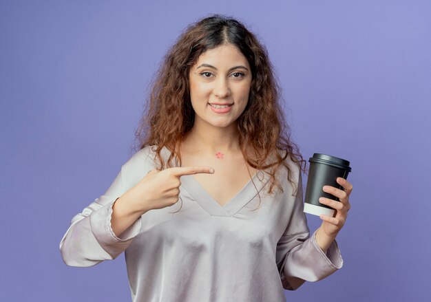 Niña bonita sonriente sosteniendo y apunta a una taza de café aislado en la pared azul