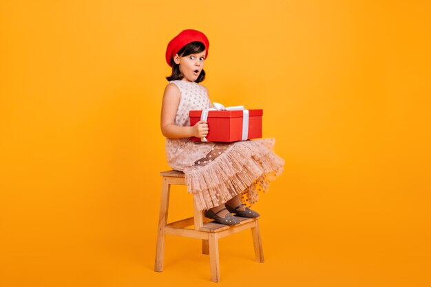 Niña bonita en vestido sentado en una silla y sosteniendo una gran caja de regalo. niño francés con regalo de cumpleaños.